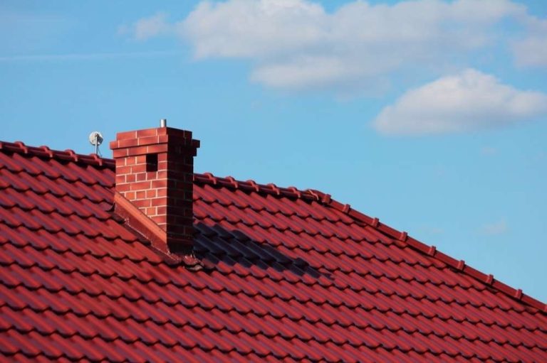 Brick chimney rising from a red tile roof under a blue sky, chimney repair for homes in Charlotte, NC by Smoke Alert